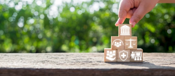 A hand places a wooden block with a government building icon on top of a pyramid of blocks featuring symbols for community, justice, healthcare, education, and support services, representing societal foundations.